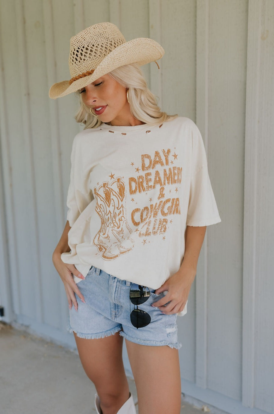 Front view of female model wearing the Day Dreamer & Cowgirl Club Distressed Graphic Tee which features Cream Cotton Fabric, Distressed Details, Round Neckline, Short Sleeves and cowgirl Boots Graphic: Day Dreamer & Cowgirl Club