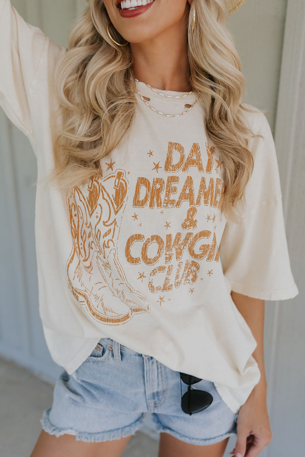 Close up view of female model wearing the Day Dreamer & Cowgirl Club Distressed Graphic Tee which features Cream Cotton Fabric, Distressed Details, Round Neckline, Short Sleeves and cowgirl Boots Graphic: Day Dreamer & Cowgirl Club