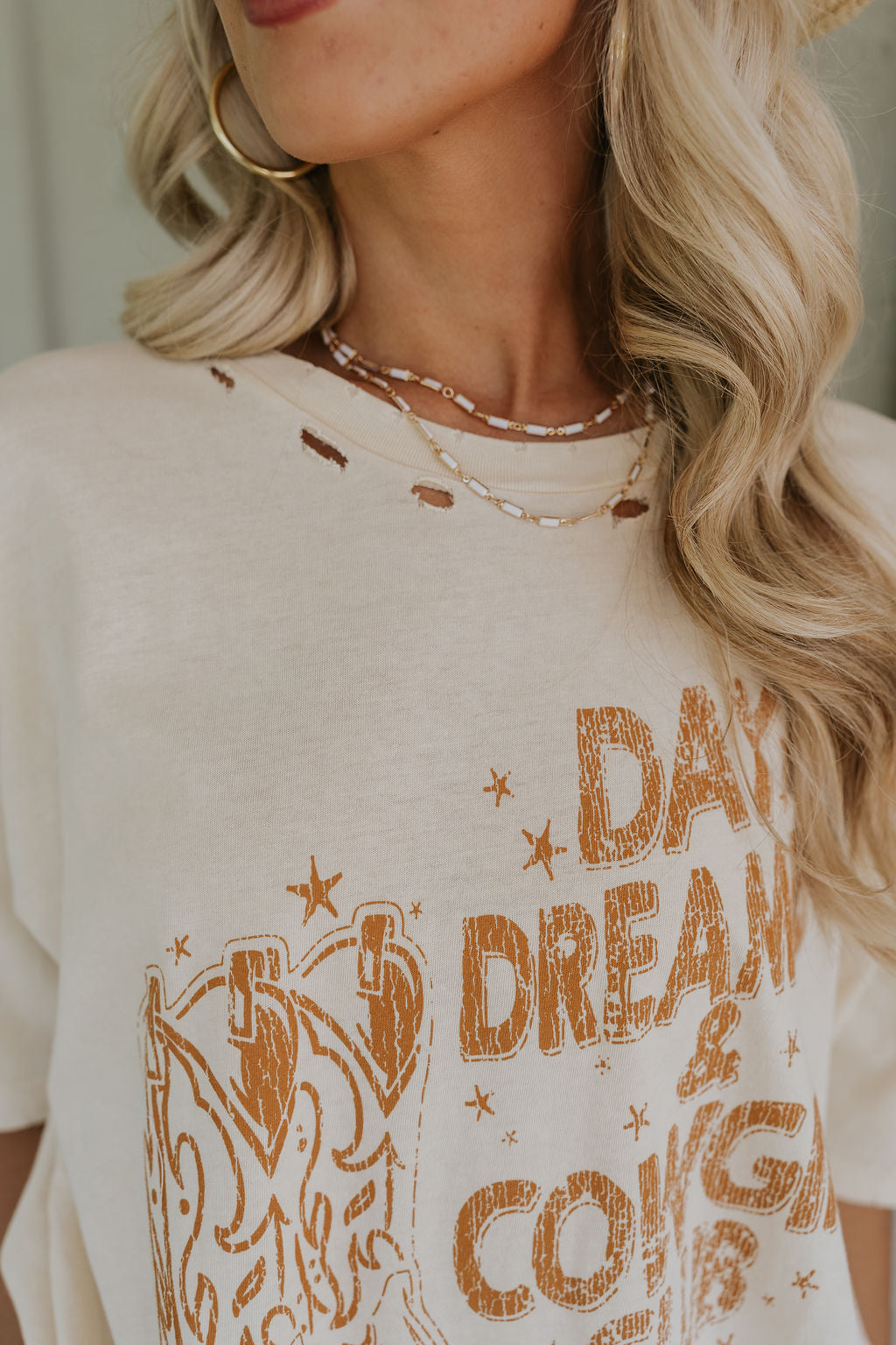 Close up view of female model wearing the Day Dreamer & Cowgirl Club Distressed Graphic Tee which features Cream Cotton Fabric, Distressed Details, Round Neckline, Short Sleeves and cowgirl Boots Graphic: Day Dreamer & Cowgirl Club