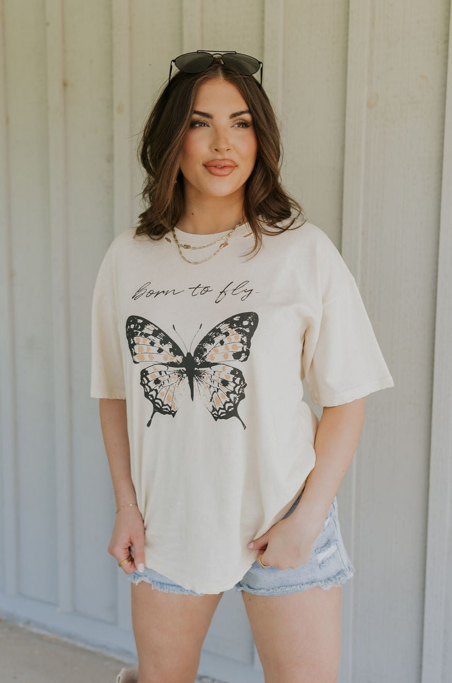 Upper body front view of female model wearing the Born To Fly Butterfly Graphic Tee that has cream fabric, a monarch butterfly graphic, "born to fly text", a round neck, oversized, and short sleeves.