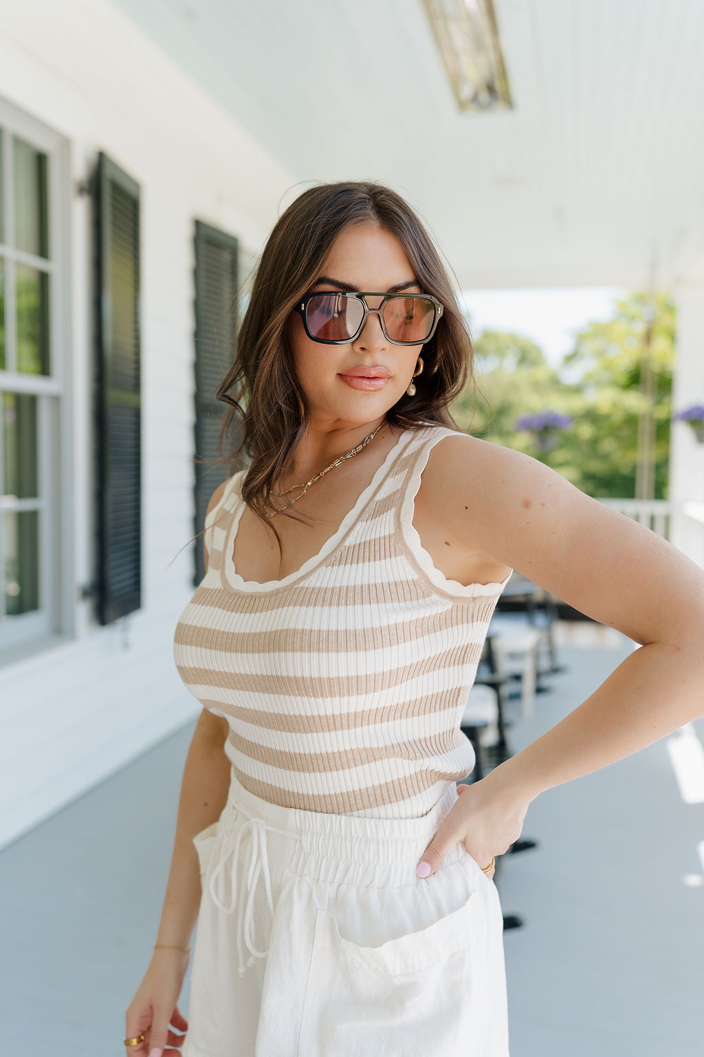 Upper body side view of brunette female model wearing the Nora Striped Tank Top in Taupe that has horizontal taupe and off white stripes, a scoop neck, scallop trim, and ribbed fabric. Top is shown tucked into shorts.