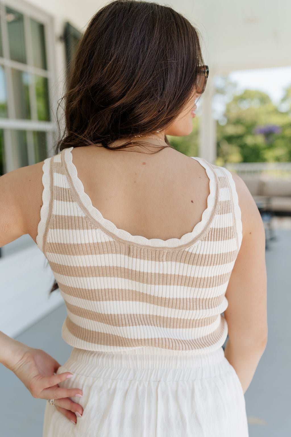 Upper body back view of brunette female model wearing the Nora Striped Tank Top in Taupe that has horizontal taupe and off white stripes, a scoop neck, scallop trim, and ribbed fabric. Top is shown tucked into shorts.