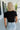 Upper body back view of blonde model wearing the Kira Short Sleeve Cropped Top in Black that has black fabric. short sleeves, a round neck, and cropped waist. Worn with denim shorts.