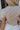 Close-up upper body back view of blonde model wearing the Kira Short Sleeve Cropped Top in Fog that has grey fabric, short sleeves, a round neck, and cropped waist. Worn with white pants.