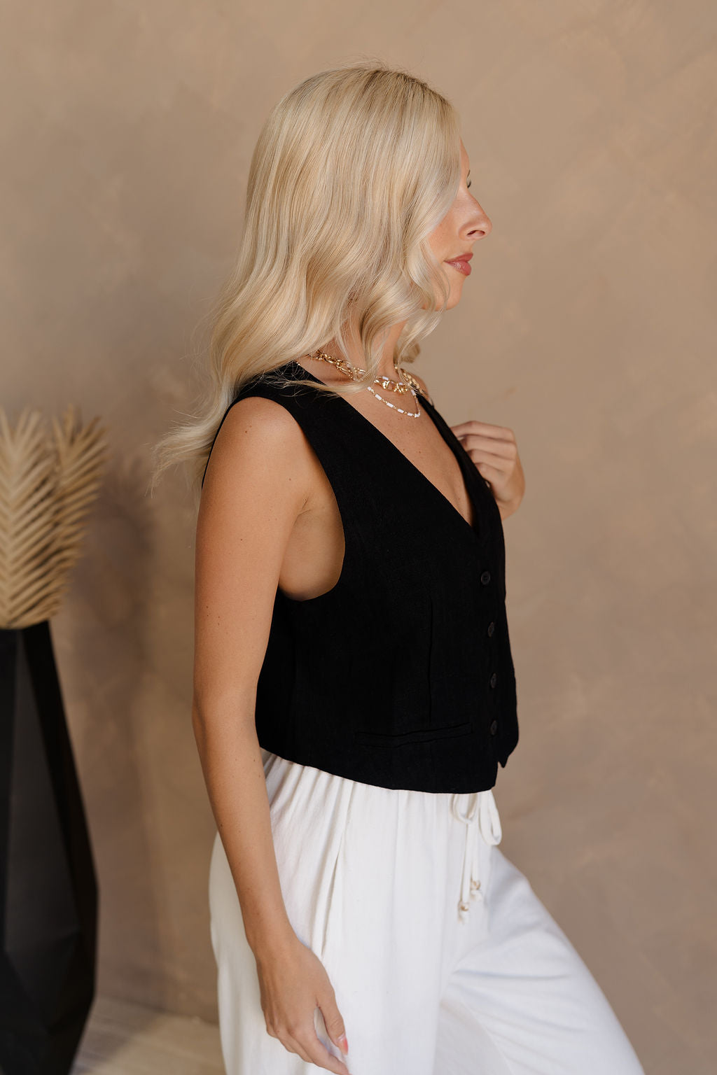 Side view of female model wearing the Jasmine Black Button-Up Vest which features Black Linen Fabric, Monochrome Button Up Front Closure, Cropped Waist, V-Neckline and Sleeveless