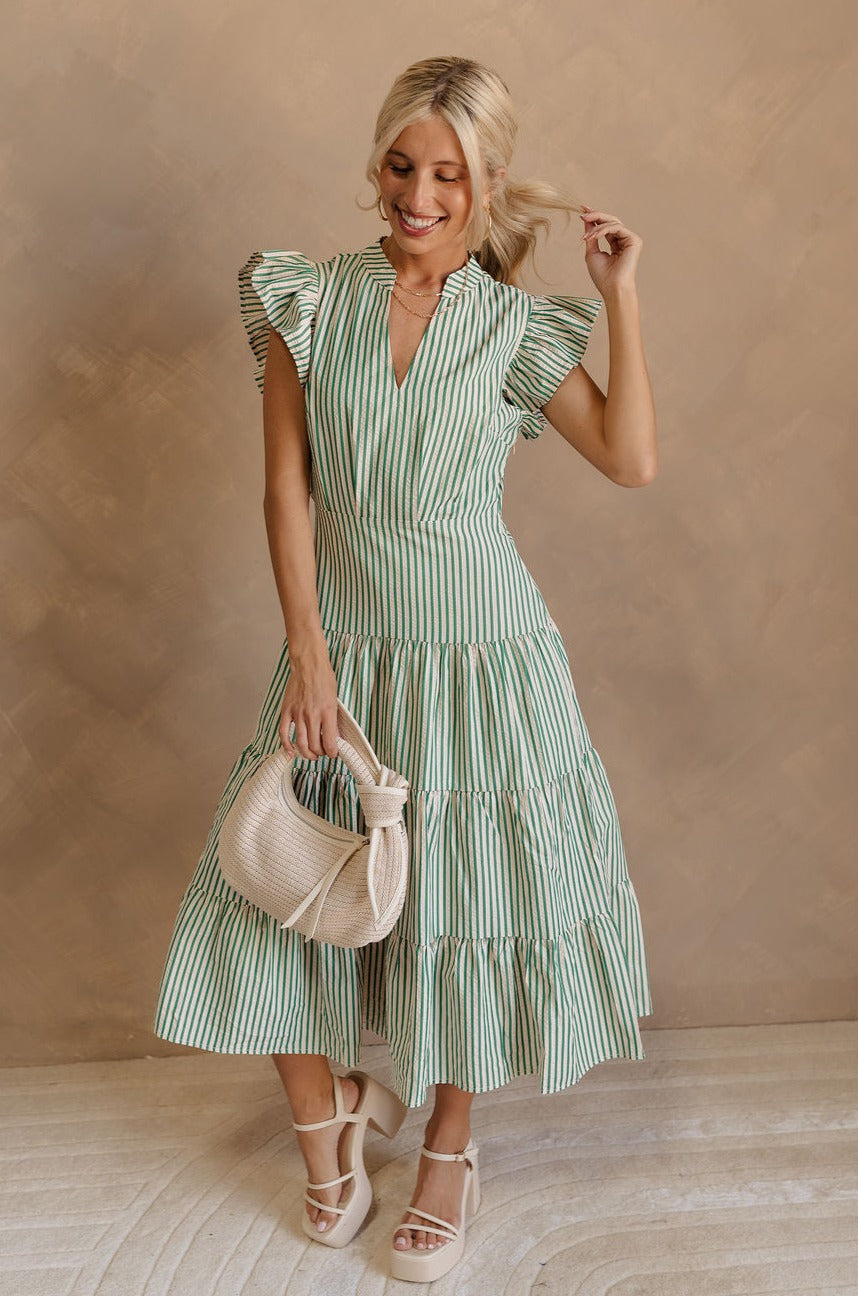 Full body front view of female model wearing the Zoey Green & Cream Striped Midi Dress that has green and cream stripes, ruffled cap sleeves, a v-neck, tiered skirt, pockets, and an open back detail. Model is holding beige purse.