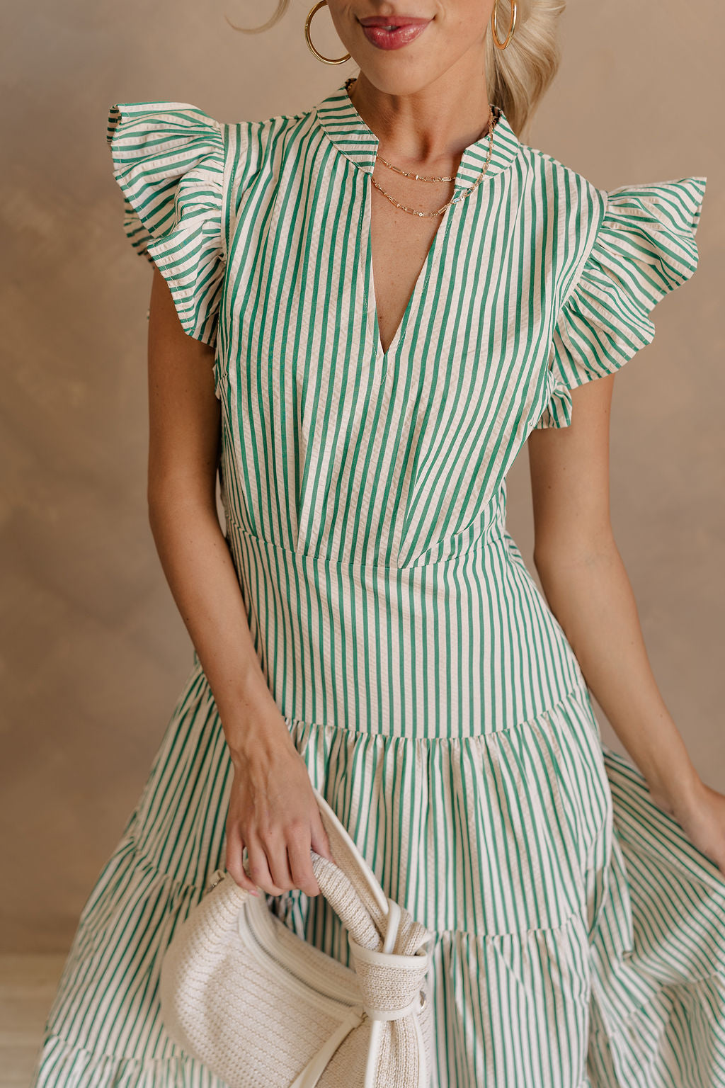 Upper body front view of female model wearing the Zoey Green & Cream Striped Midi Dress that has green and cream stripes, ruffled cap sleeves, a v-neck, tiered skirt, pockets, and an open back detail. Model is holding beige purse.