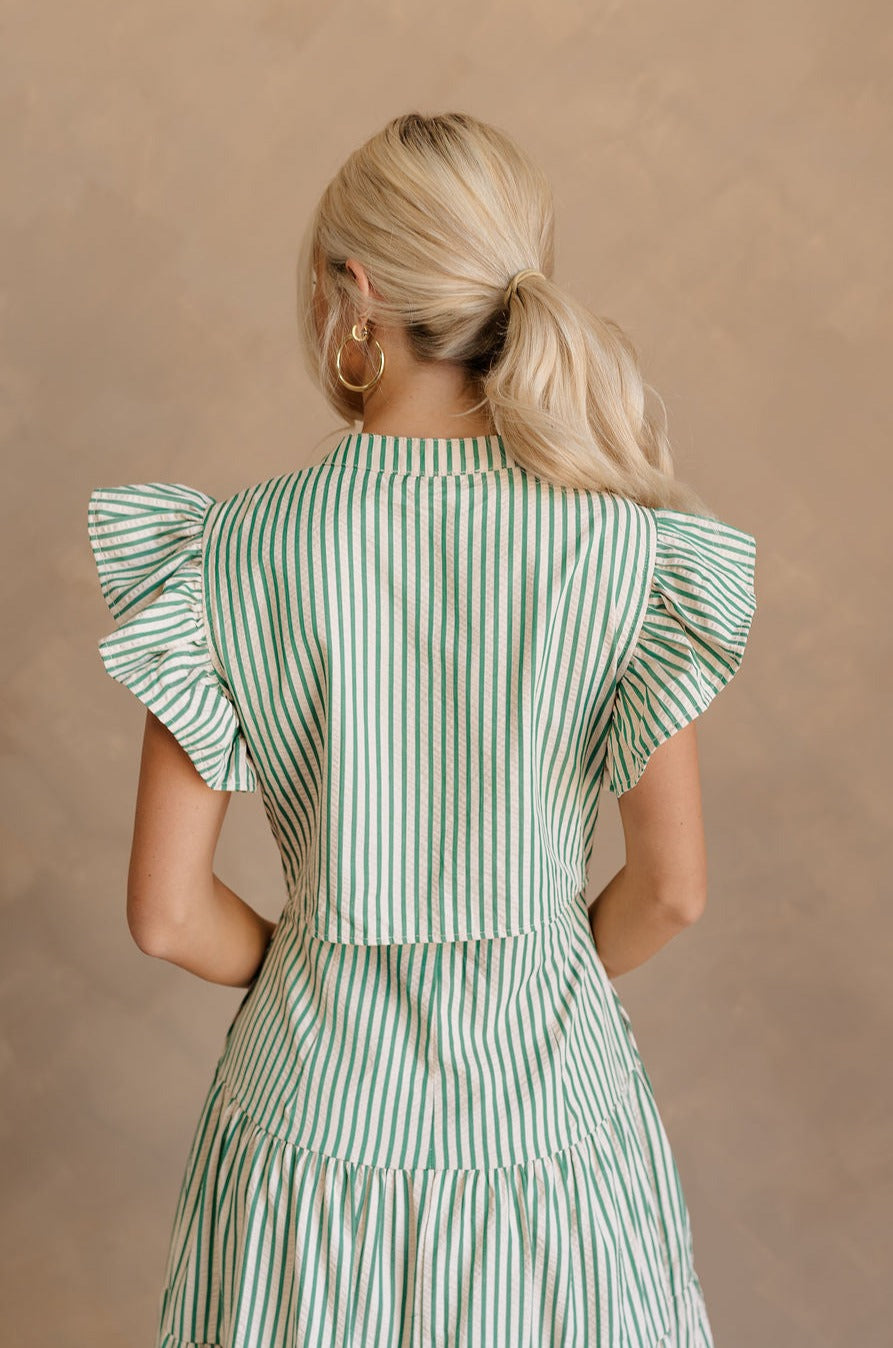 Upper body back view of female model wearing the Zoey Green & Cream Striped Midi Dress that has green and cream stripes, ruffled cap sleeves, a v-neck, tiered skirt, pockets, and an open back detail.