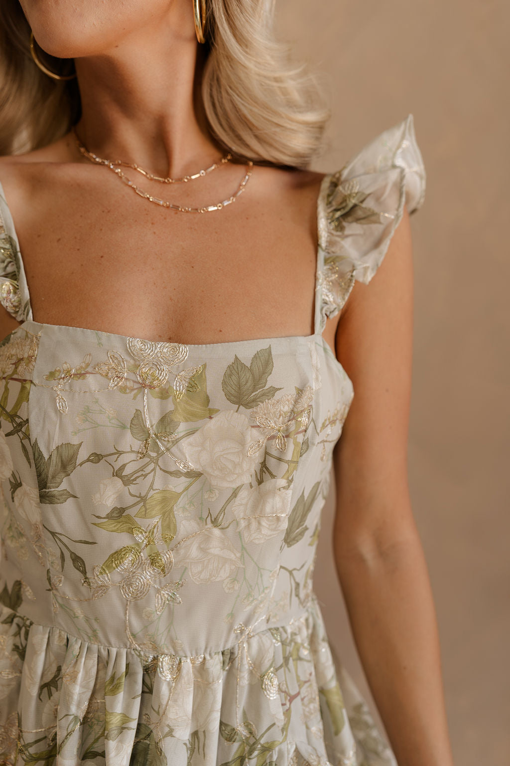 Close up view of female model wearing the Thea Green & Gold Floral Ruffle Maxi Dress which features Green sheer fabric with green & beige floral print, Ivory & Gold embroidery, Ruffled adjustable straps, Side slit with ruffle trim, Beige lining and Back zipper and hook closure