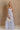 Full body view of female model wearing the Cali Purple Floral Maxi Dress which features Sheer purple fabric, Purple floral print and white floral embroidery, Purple lining, Bustier top, Adjustable straps, Open back with a tie closure and elastic waist and Tiered skirt