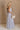 Full body side view of female model wearing the Cali Purple Floral Maxi Dress which features Sheer purple fabric, Purple floral print and white floral embroidery, Purple lining, Bustier top, Adjustable straps, Open back with a tie closure and elastic waist and Tiered skirt