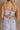 Close up back view of female model wearing the Cali Purple Floral Maxi Dress which features Sheer purple fabric, Purple floral print and white floral embroidery, Purple lining, Bustier top, Adjustable straps, Open back with a tie closure and elastic waist and Tiered skirt