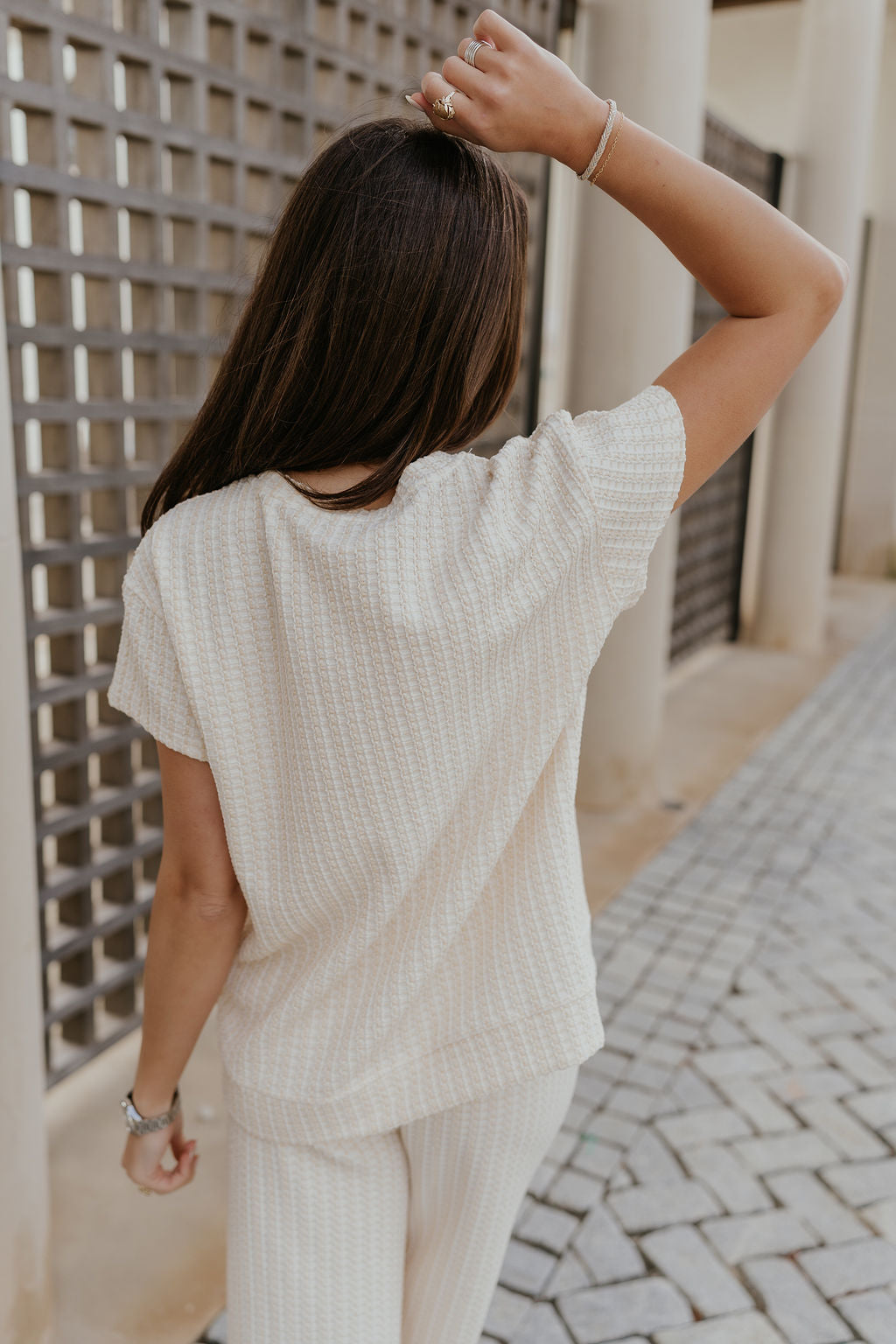 Back view of female model wearing the Willow Cream Woven Short Sleeve Top which features Cream and White Woven Design, Thick Hem, Round Neckline and Short Sleeves