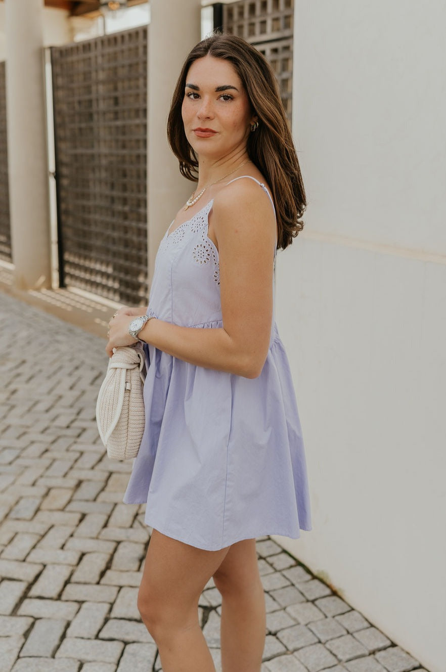 Side view of female model wearing the Lucia Lavender Eyelet Sleeveless Mini Dress which features Lavender Lightweight Fabric, Lavender Lining, Mini Length, Eyelet Pattern, Scalloped Upper Hem and Adjustable Straps