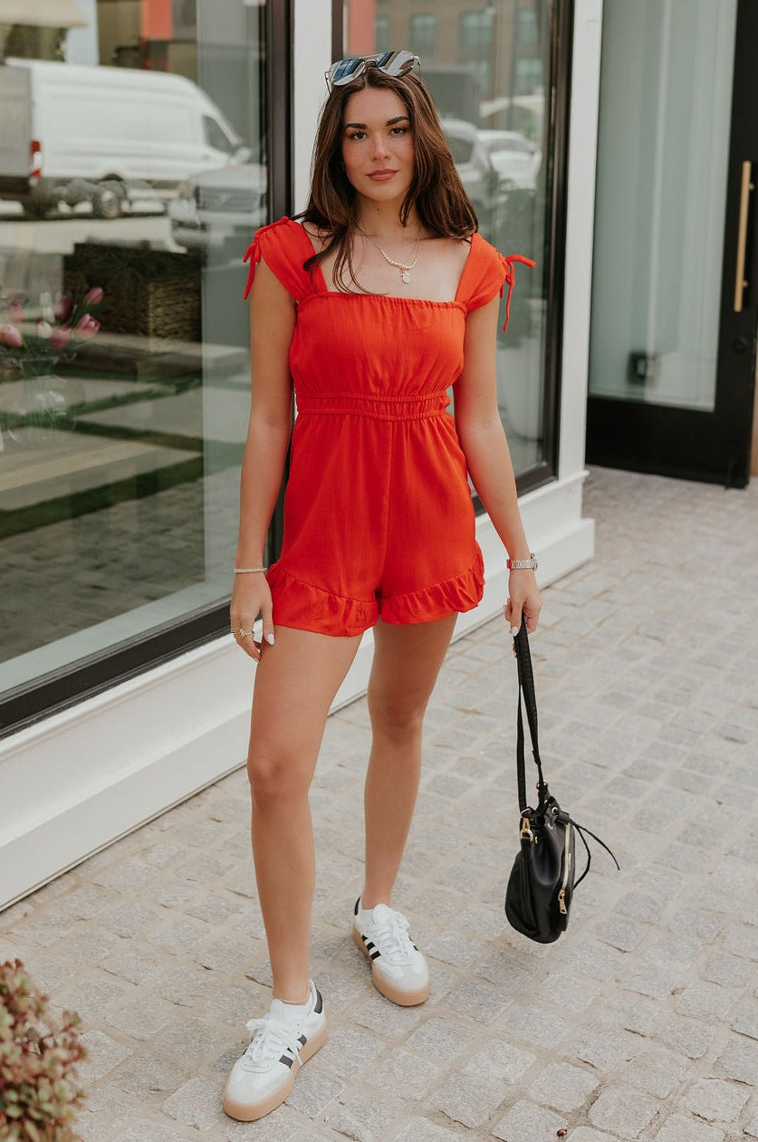 Full body view of female model wearing the Rosalie Red Sleeveless Romper which features Tomato Red Lightweight Fabric, Ruffle Hem, Elastic Waistband, Square Neckline and Thick Straps with Tie Details