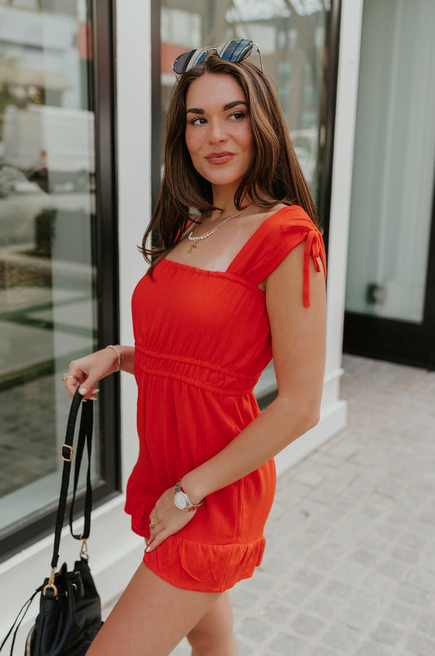 Side view of female model wearing the Rosalie Red Sleeveless Romper which features Tomato Red Lightweight Fabric, Ruffle Hem, Elastic Waistband, Square Neckline and Thick Straps with Tie Details