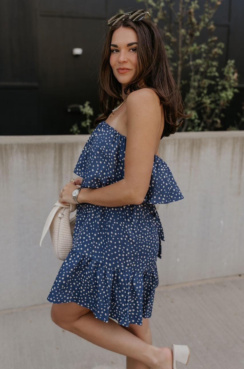 Side view of female model wearing the Lena Navy & White Polka Dot Strapless Mini Dress which features Navy and Cream Polka Dot Design, Ruffle Hem Skirt, Upper Ruffle Detail and Strapless
