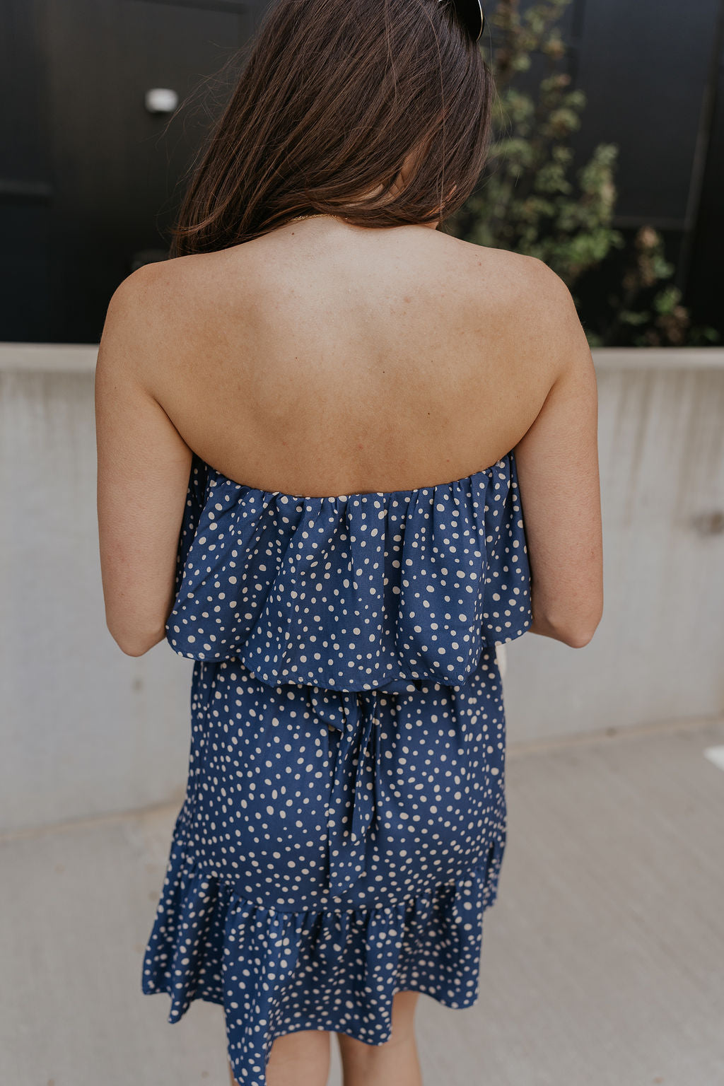 Back view of female model wearing the Lena Navy & White Polka Dot Strapless Mini Dress which features Navy and Cream Polka Dot Design, Ruffle Hem Skirt, Upper Ruffle Detail and Strapless