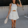 Full body front view of female model wearing the Roselyn White Tiered Mini Skirt that has white fabric, two tiers, and an elastic waist. Worn with white tank top.