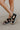 Side view of female model wearing the Mackenna Sandal in Black which features smooth, sleak black leather upper, white rugged sole, adjustable buckle, crisscross straps, cushioned footbed and back zipper with leather pull