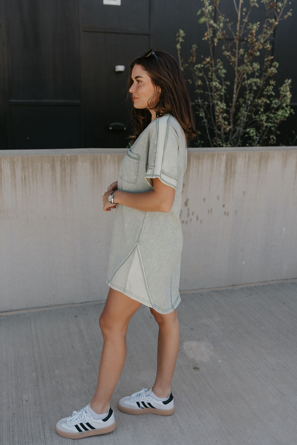Full body side view of female model wearing the Dalia Sage Green Knit Mini Dress that has washed sage green fabric, a front pocket, and short sleeves.