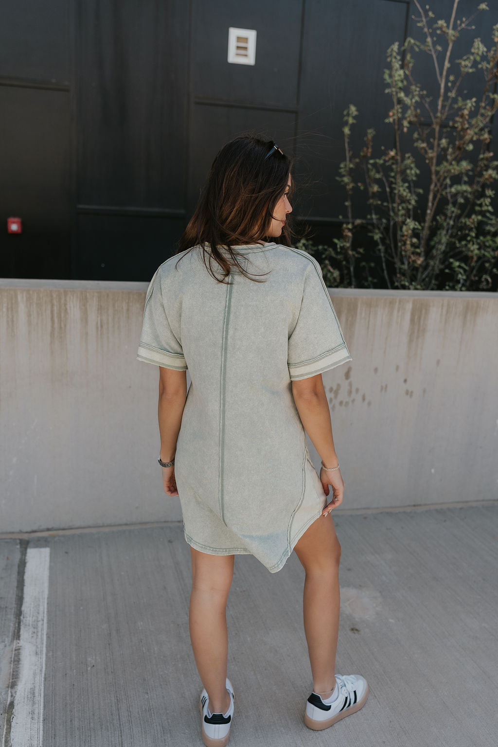 Full body back view of female model wearing the Dalia Sage Green Knit Mini Dress that has washed sage green fabric, a front pocket, and short sleeves.
