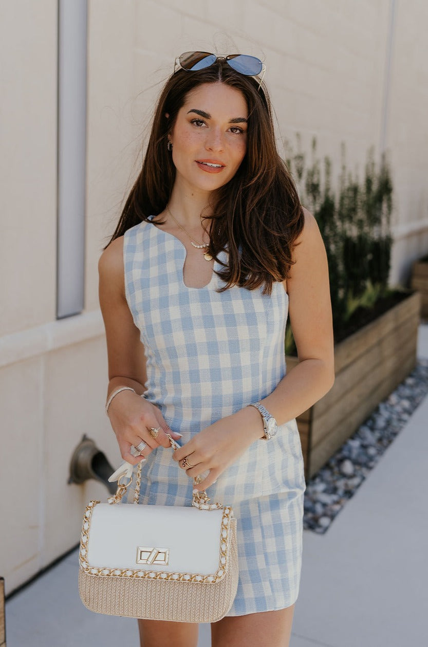 Upper body front view of female model wearing the Georgina Blue Gingham Mini Dress that has light blue and white gingham fabric, a noticed neckline, a side slit, and is sleeveless. Model is holding purse in front of her