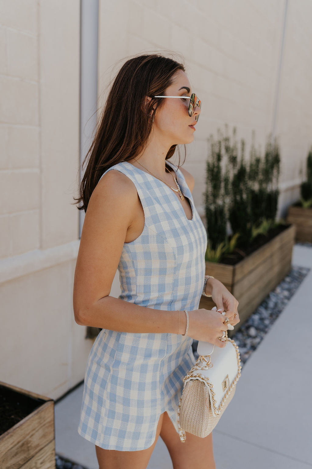 Upper body side view of female model wearing the Georgina Blue Gingham Mini Dress that has light blue and white gingham fabric, a noticed neckline, a side slit, and is sleeveless. Model is holding purse in front of her