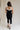 Full body back view of female model wearing the Marissa Black & White Trim Midi Dress which features Black Lightweight Fabric, Midi Length, Black Lining, White Trim Details, Back Slit , Square Necklineand Side Monochrome Zipper with Hook Closure