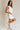 Full body side view of female model wearing the Taylor Off White Bow Cut-Out Mini Dress which features White Lightweight Fabric, White Lining, Mini Length, Square Neckline, Side Zipper with Hook Closure and Bow Cut-Out Back Detail