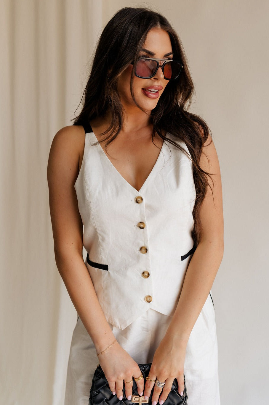 front view of female model wearing the Amelia Off White & Black Button-Up Vest which features White Linen Fabric, Black Trim Details, Front Pockets, Brown Tortoise Button Up, V-Neckline and Sleeveless
