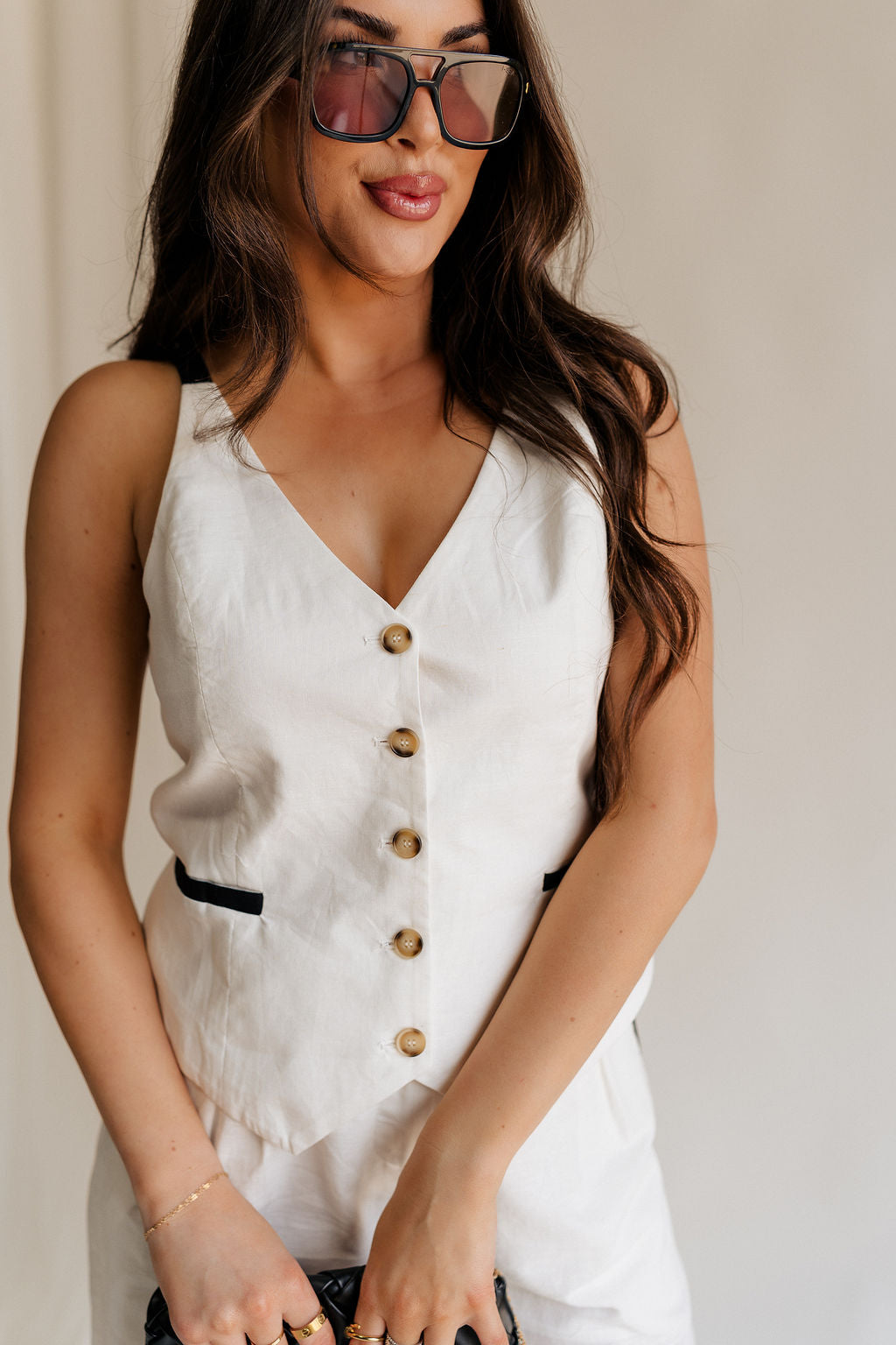 close up view of female model wearing the Amelia Off White & Black Button-Up Vest which features White Linen Fabric, Black Trim Details, Front Pockets, Brown Tortoise Button Up, V-Neckline and Sleeveless