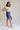 Full body side view of model wearing the Mara Blue High Waist Shorts that have blue fabric, a high-rise elastic back waist. Worn with beige top.