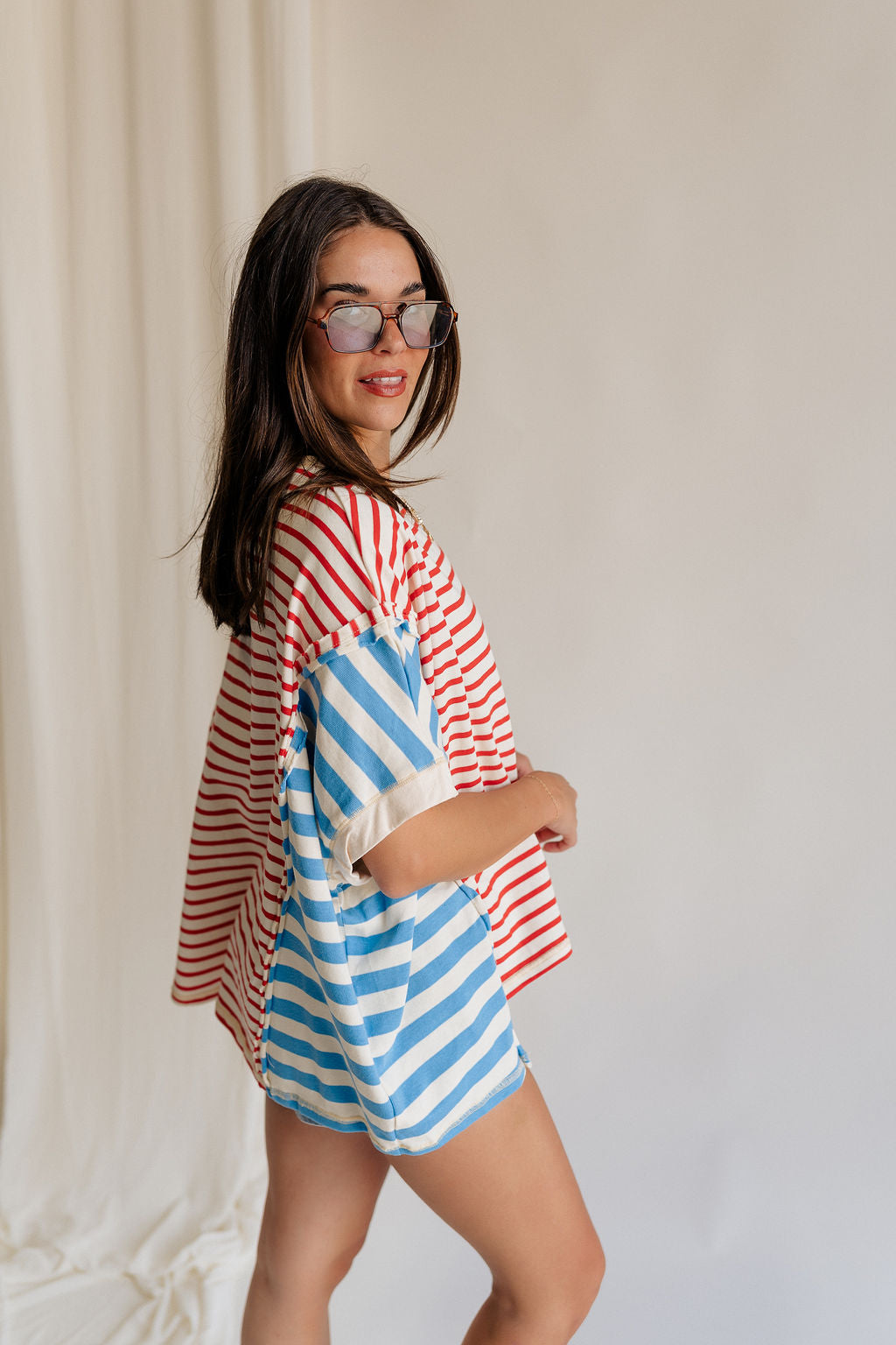 Side view of female model wearing the Abigail Red, Cream, & Blue Striped Top that has a red striped center, blue striped sides, a round neck, short sleeves, and oversize fit.