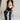 Full body front view of female model wearing the Remi Rhinestone Slit Black Jeans that have black denim, thigh slits with rhinestone trim, and pockets. Model is sitting on stool.