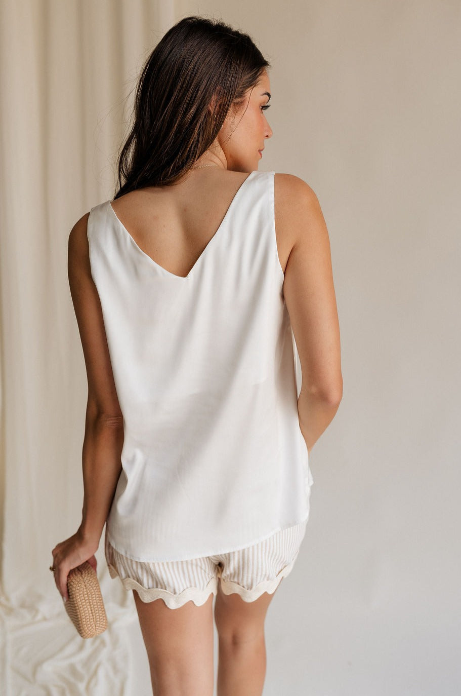 Back view of female model wearing the Monroe Off White Satin Tank Top that has off white satin fabric and a v-neckline.