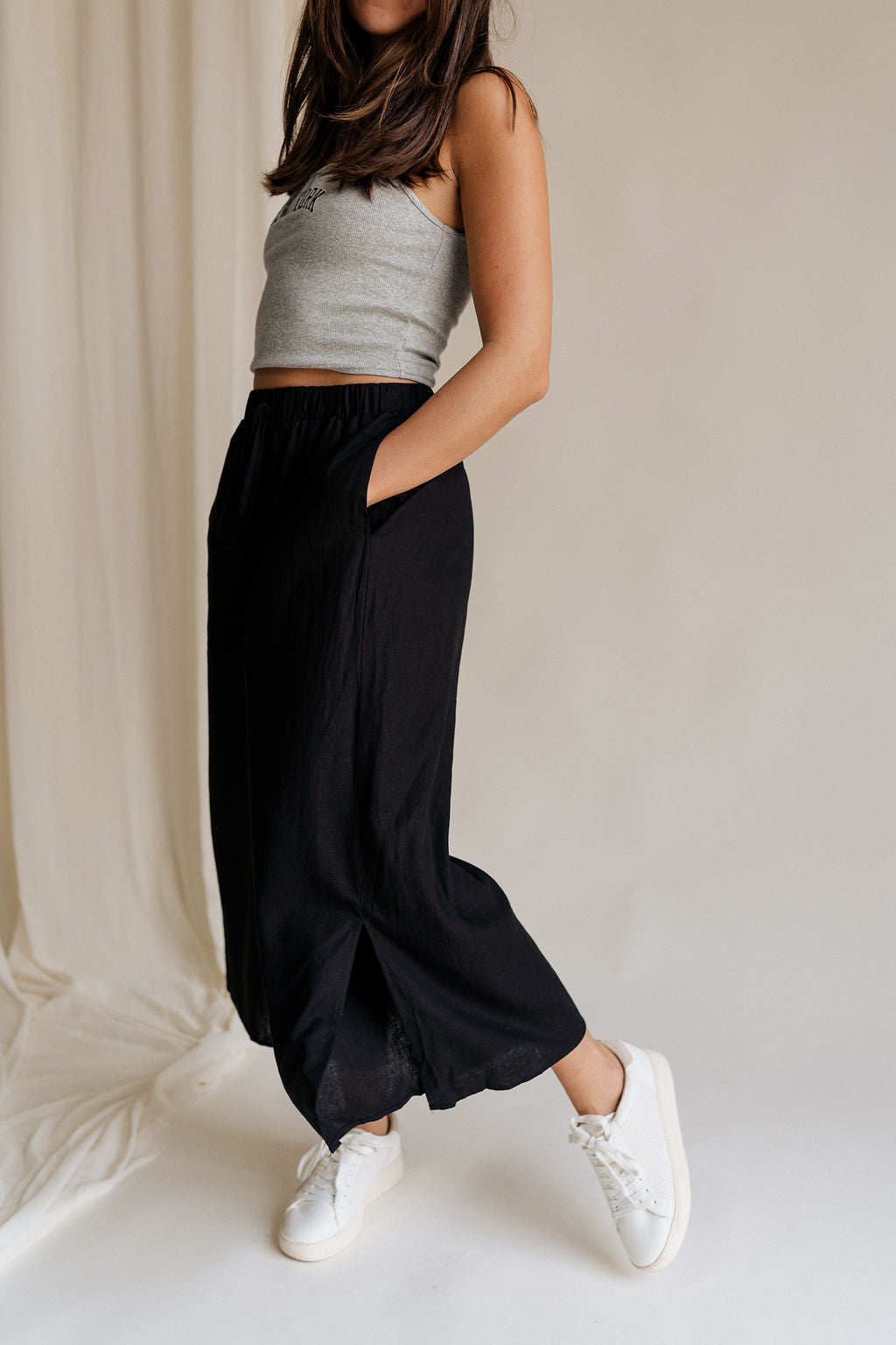 Frontal side view of female model wearing the Meredith Black Cropped Wide Leg Pants that have black fabric, cropped wide legs, side slits, and a drawstring waist.