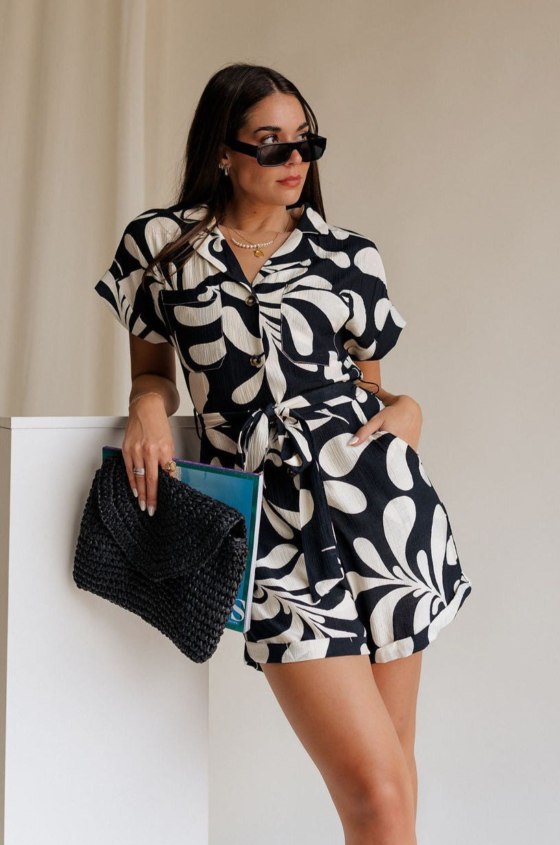 Front view of model wearing the Melanie Black & Cream Printed Romper that has black and cream fabric with a swirl print, a button up front, collar, tie belt, and short sleeves. 