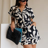 Front view of model wearing the Melanie Black & Cream Printed Romper that has black and cream fabric with a swirl print, a button up front, collar, tie belt, and short sleeves. 