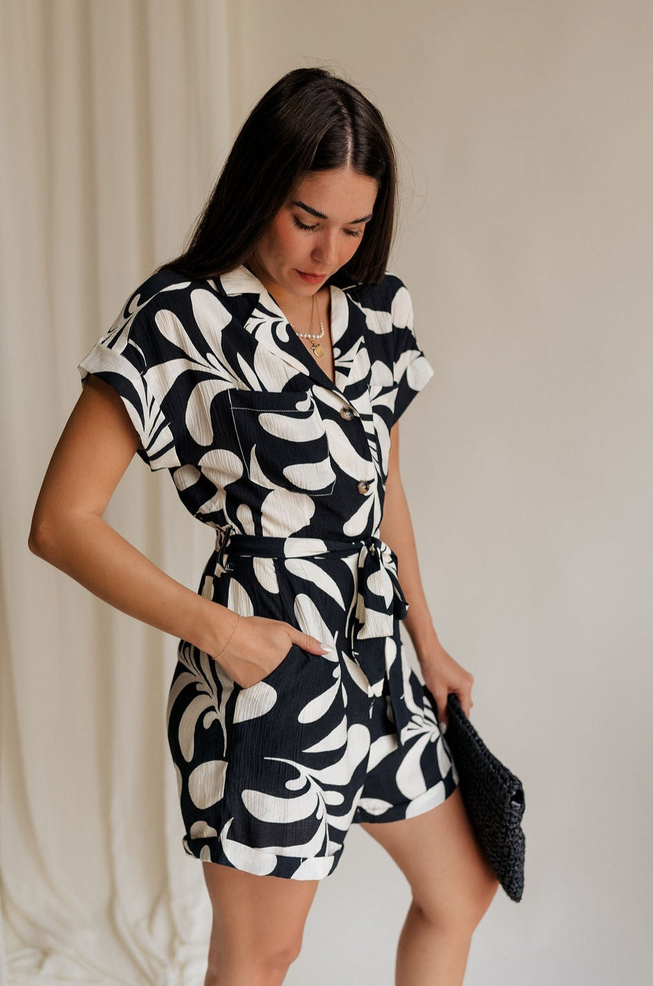 Frontal side view of model wearing the Melanie Black & Cream Printed Romper that has black and cream fabric with a swirl print, a button up front, collar, tie belt, and short sleeves. 
