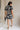Full body back view of model wearing the Melanie Black & Cream Printed Romper that has black and cream fabric with a swirl print, a button up front, collar, tie belt, and short sleeves. 