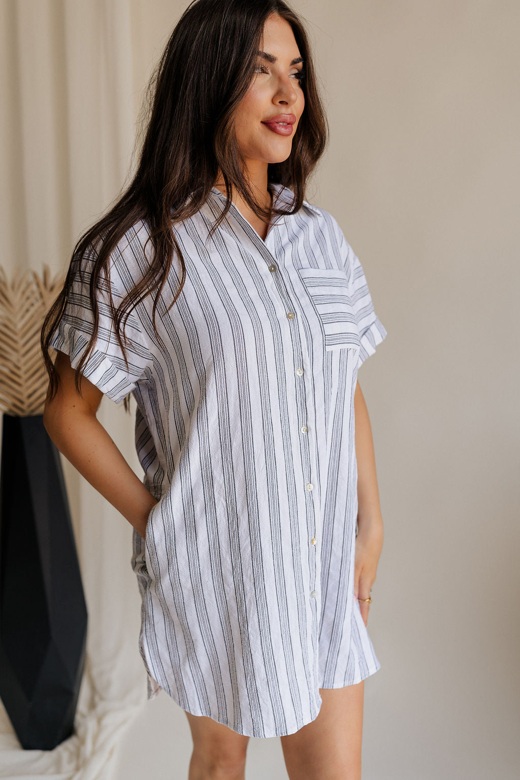 Frontal side view of female model wearing the Jenna White & Black Striped Mini Dress that has white fabric with vertical black stripes, a front pocket, button up front, short sleeves, and collar.