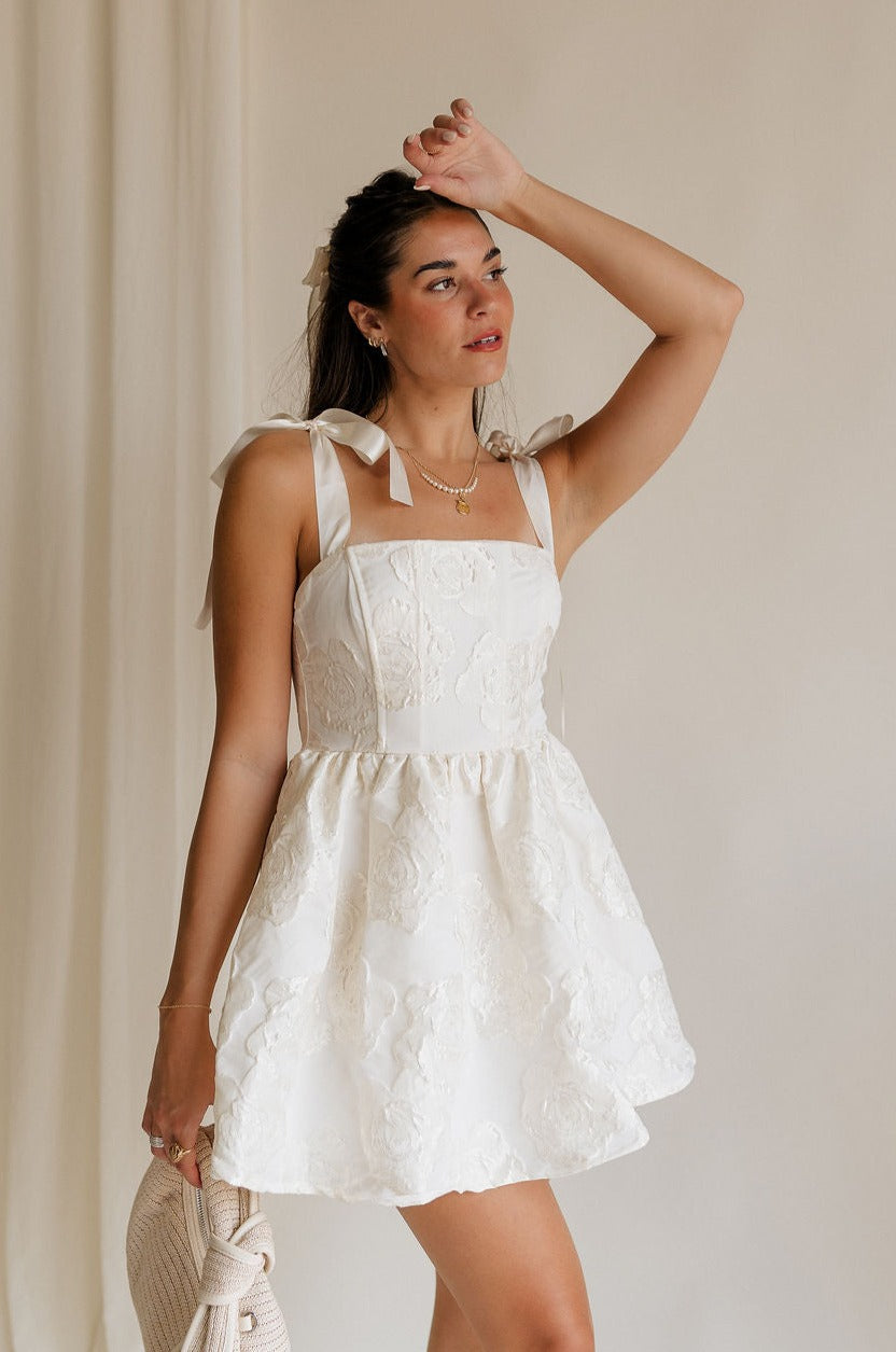 Front view of female model wearing the Sasha Off White Tie Strap Mini Dress that has cream fabric with rosette florals, cream tie straps, a straight neckline, and fit and flare style.