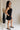 Full body side view of female model wearing the Thea Black Square Neckline Mini Dress which features Black Linen Fabric, Black Lining, Mini Length, Side Pockets, Square Neckline, Sleeveless and Monochrome Back Zipper with Hook Closure