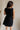 back view of female model wearing the Thea Black Square Neckline Mini Dress which features Black Linen Fabric, Black Lining, Mini Length, Side Pockets, Square Neckline, Sleeveless and Monochrome Back Zipper with Hook Closure