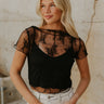 Upper body front view of female model wearing the Vanessa Cropped Lace Top in Black that has a short sleeve sheer lace top over an ivory cami. Worn with white denim skirt.