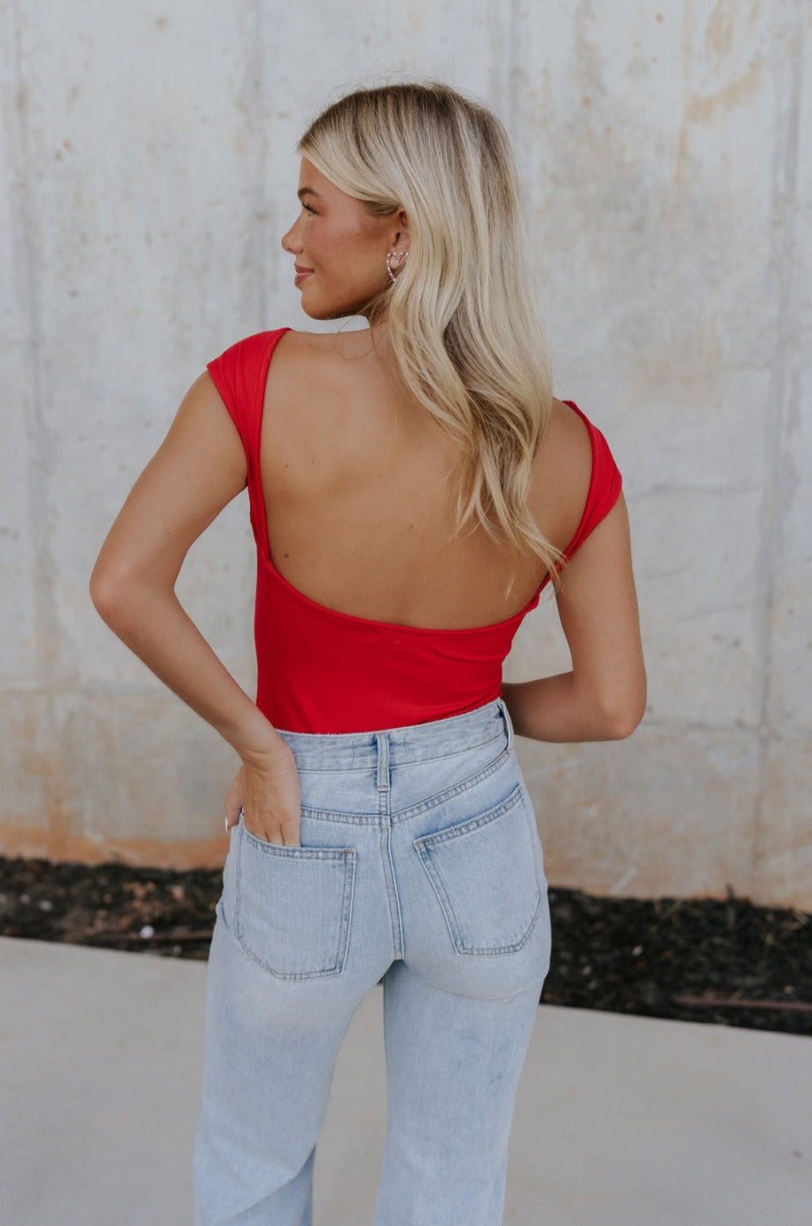 Back view of female model wearing the Ryleigh Red Backless Bodysuit that has red knit fabric, an open back, and a scoop neckline. Worn with jeans.