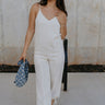 Full body front view of female model wearing the Parker Beige Linen Jogger Jumpsuit that has beige linen fabric, a v-neck, thin straps, pockets, and jogger style legs with ankle ties.