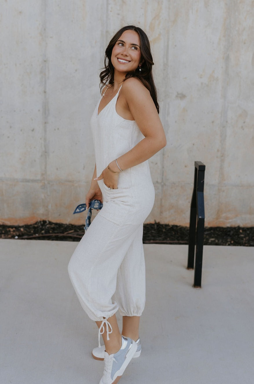 Full body side view of female model wearing the Parker Beige Linen Jogger Jumpsuit that has beige linen fabric, a v-neck, thin straps, pockets, and jogger style legs with ankle ties.
