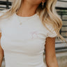 Close-up front view of model wearing the Lana Embroidered Bow Cream Cropped Tee that has cream ribbed fabric, a pink embroidered bow on the chest, and short sleeves.